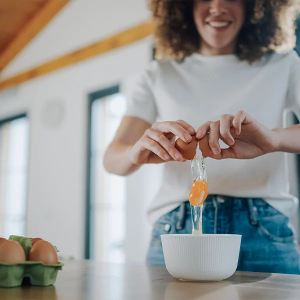 A person cracks an egg into a bowl on a kitchen counter, with a carton of eggs nearby.