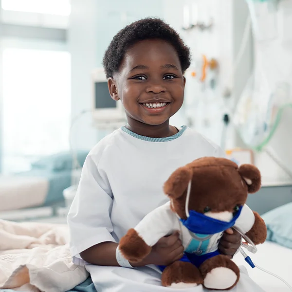 A smiling child in a hospital gown sits on a bed holding a teddy bear dressed with a face mask and medical tubing in a hospital room.
