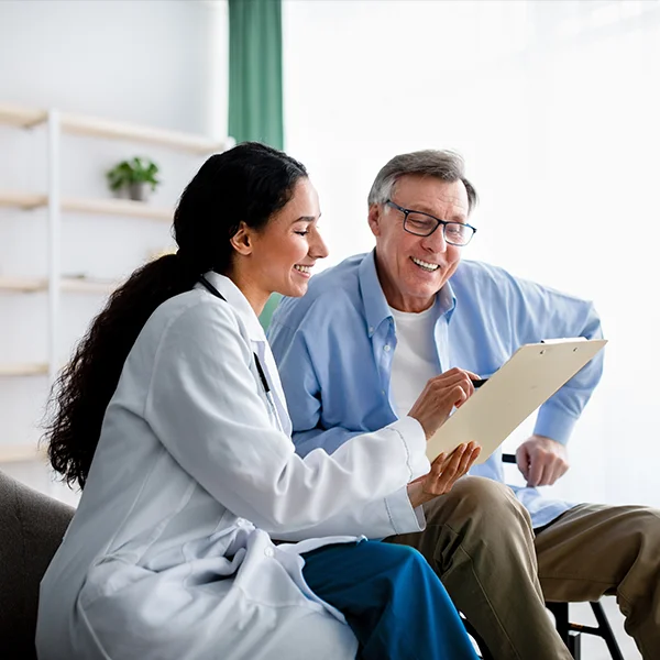 A female doctor and an older male patient sit together, smiling as they review information on a clipboard in a bright, modern exam room.