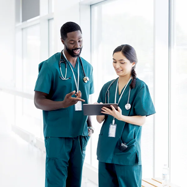 Two healthcare professionals in scrubs stand in a bright hospital corridor, smiling and reviewing information together on a tablet.