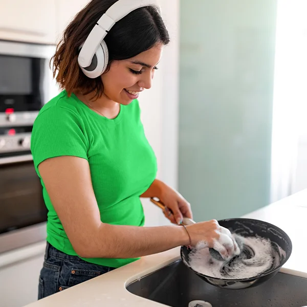 A woman wearing headphones smiles while washing a pan with soapy water at a kitchen sink.