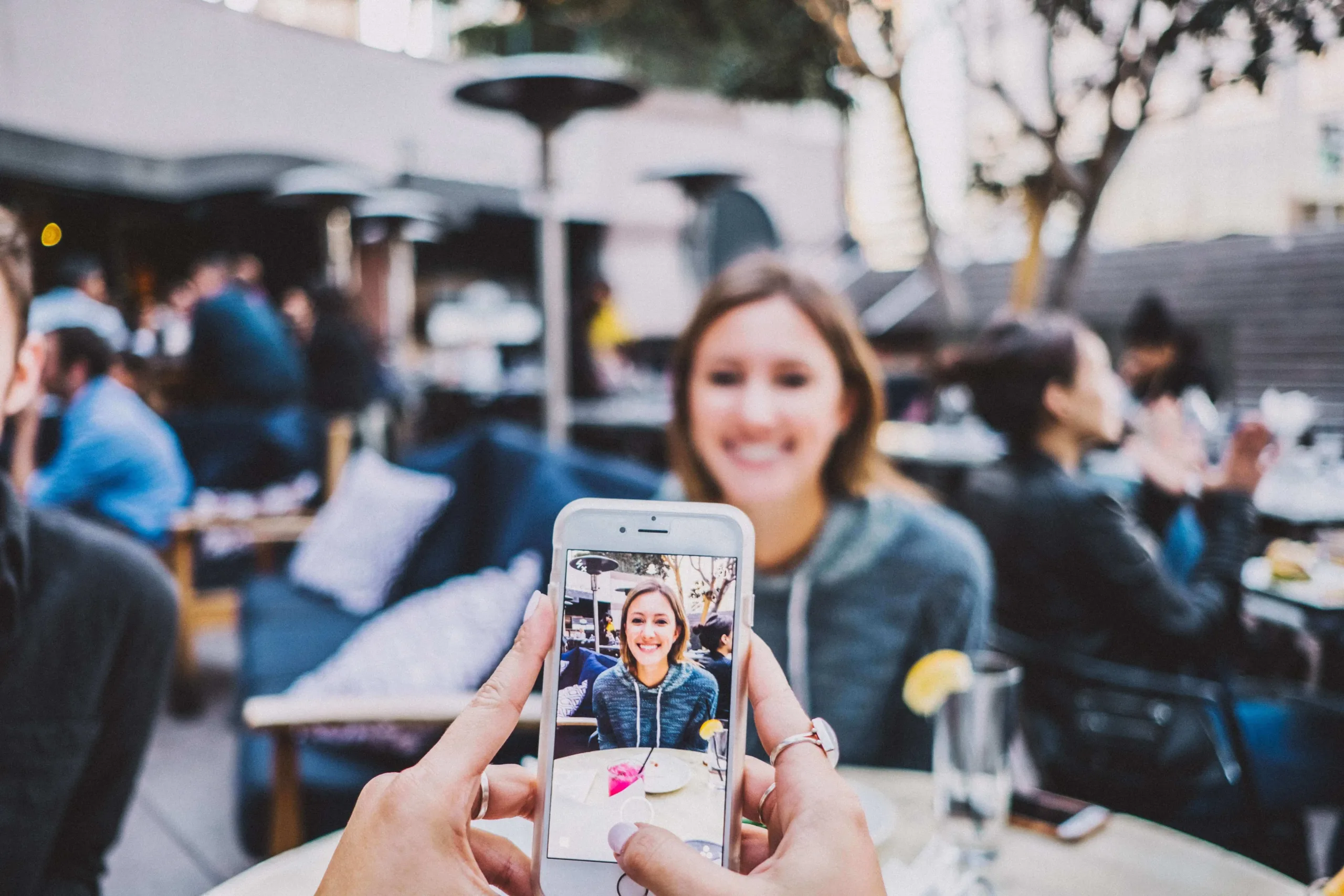 Someone photographing a smiling person at an outdoor cafe using a smartphone camera, with the subject visible both in real life and on the phone screen