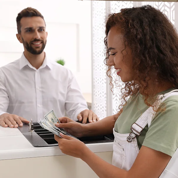 A woman counts cash at a bank counter while a teller stands behind the desk smiling.