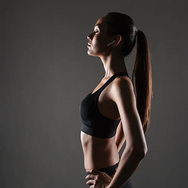 A woman in athletic wear stands in profile with her eyes closed, wearing wireless earbuds against a dark background.