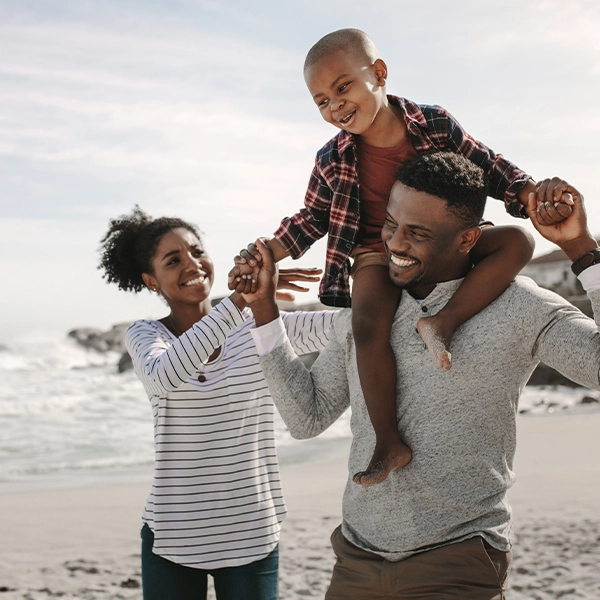 A family enjoys time together on a beach, with a man carrying a smiling child on his shoulders while a woman holds the child’s hand nearby.