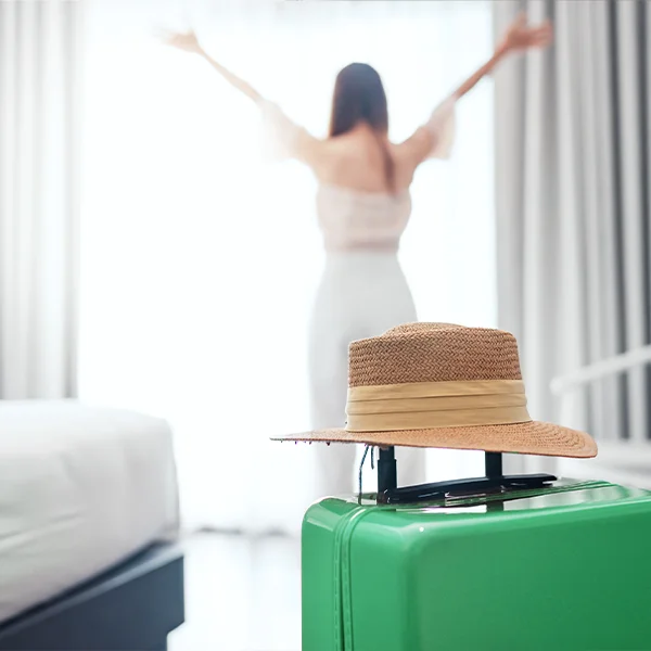 A straw hat rests on a green suitcase in a hotel room while a person in the background stretches near a bright window.