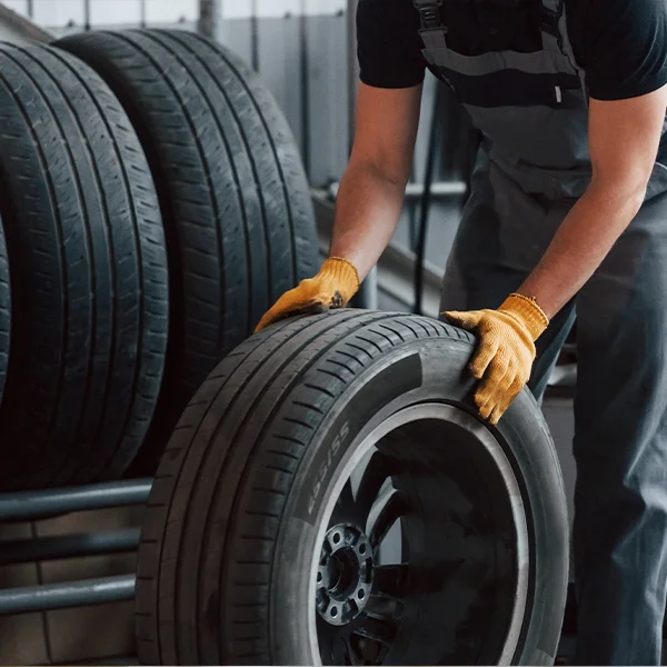 A mechanic wearing gloves lifts a car tire in a workshop with stacked tires in the background.