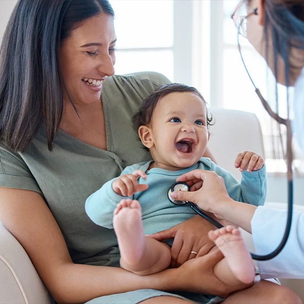 A smiling mother holds her baby while a doctor uses a stethoscope to examine the child.