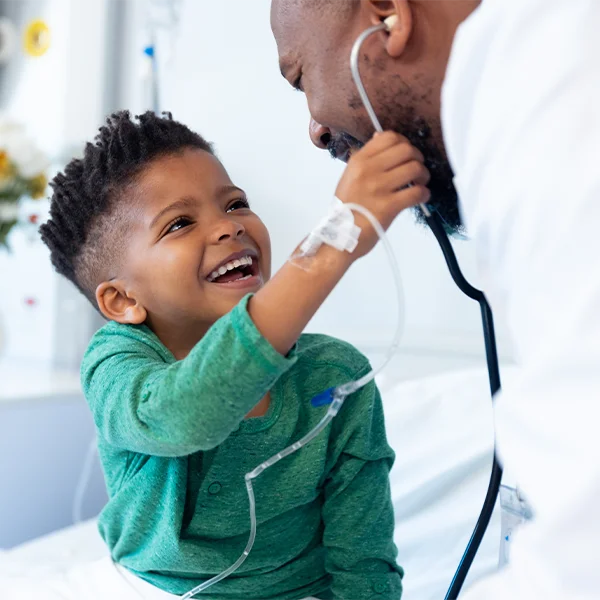 A young child in a hospital bed smiles while playfully holding a doctor’s stethoscope as the doctor leans in.