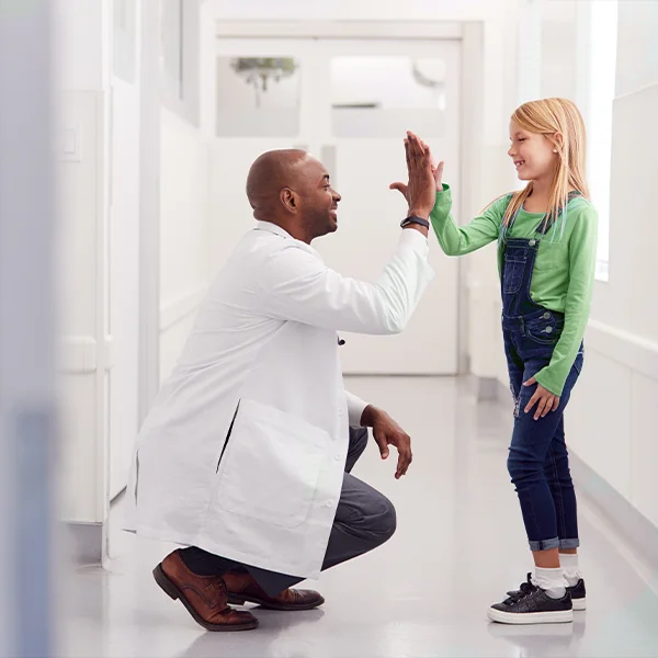 A doctor kneels in a hallway to give a young girl a high-five as she smiles.