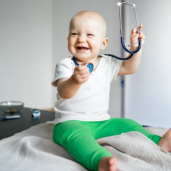 A smiling baby sitting on an exam table holds up a stethoscope, appearing playful and curious in a medical setting.