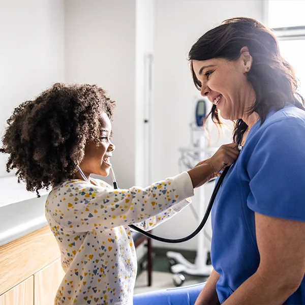 A young child is playfully using a stethoscope to listen to a smiling female healthcare worker’s heartbeat in a bright medical exam room.