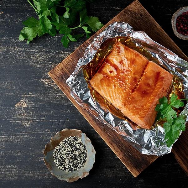 A cooked salmon fillet rests on foil atop a wooden board, surrounded by herbs and seasonings.