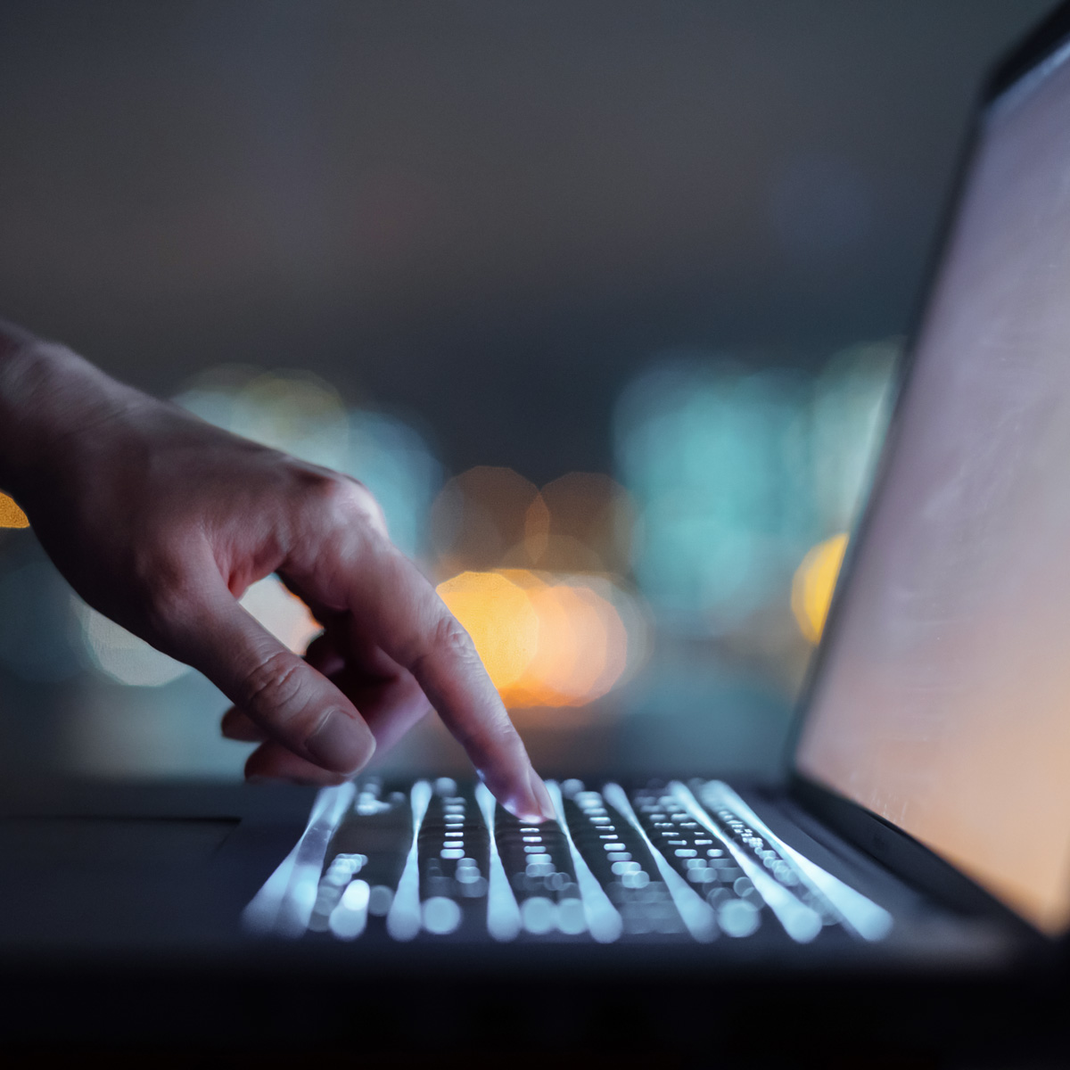 A close-up of a hand typing on a laptop keyboard at night, with a softly lit screen and blurred city lights in the background.