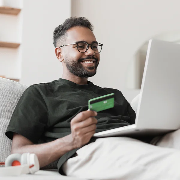 A man sits on a couch using a laptop while holding a credit card, smiling as he shops online.