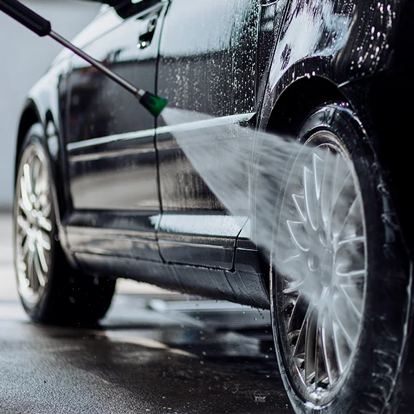 A car is being washed with a high-pressure hose, spraying water onto the wheel and side panel.