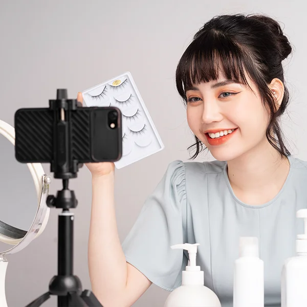 A woman smiles while holding a set of false eyelashes and filming herself with a smartphone on a tripod, surrounded by beauty products.