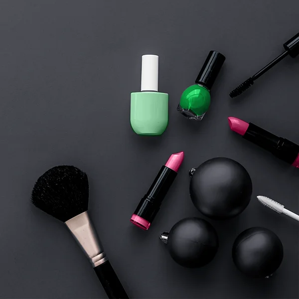 An assortment of makeup products, including lipstick, nail polish, mascara, and a brush, arranged on a dark surface.