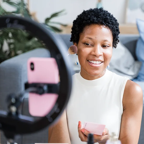 A woman smiles while applying makeup and filming herself with a smartphone inside a ring light.