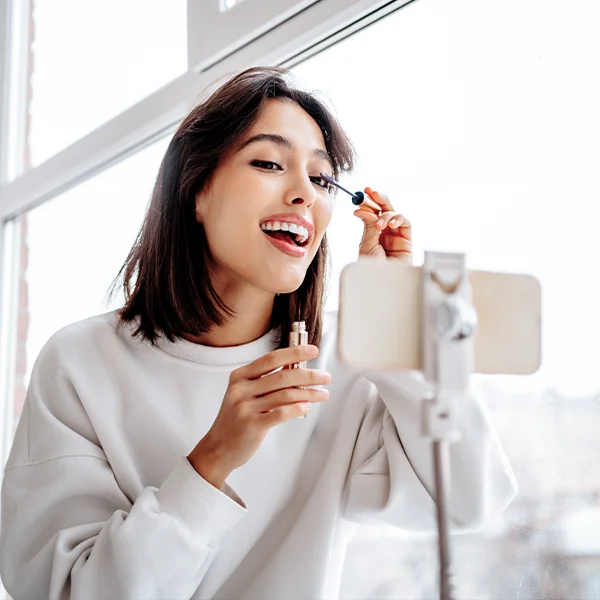 A woman applies mascara while looking into a smartphone mounted on a stand near a window.
