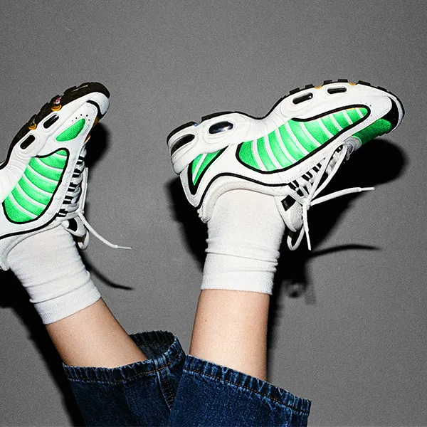 A person wearing white sneakers with neon green accents and white socks lifts their feet against a plain gray background.