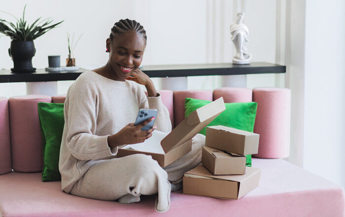 Woman sitting on a couch with packages stacked next to her which she smiles at her phone.