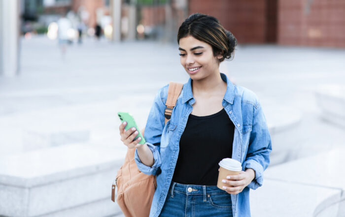 A woman looking at their cell phone with a coffee cup in their opposite hand