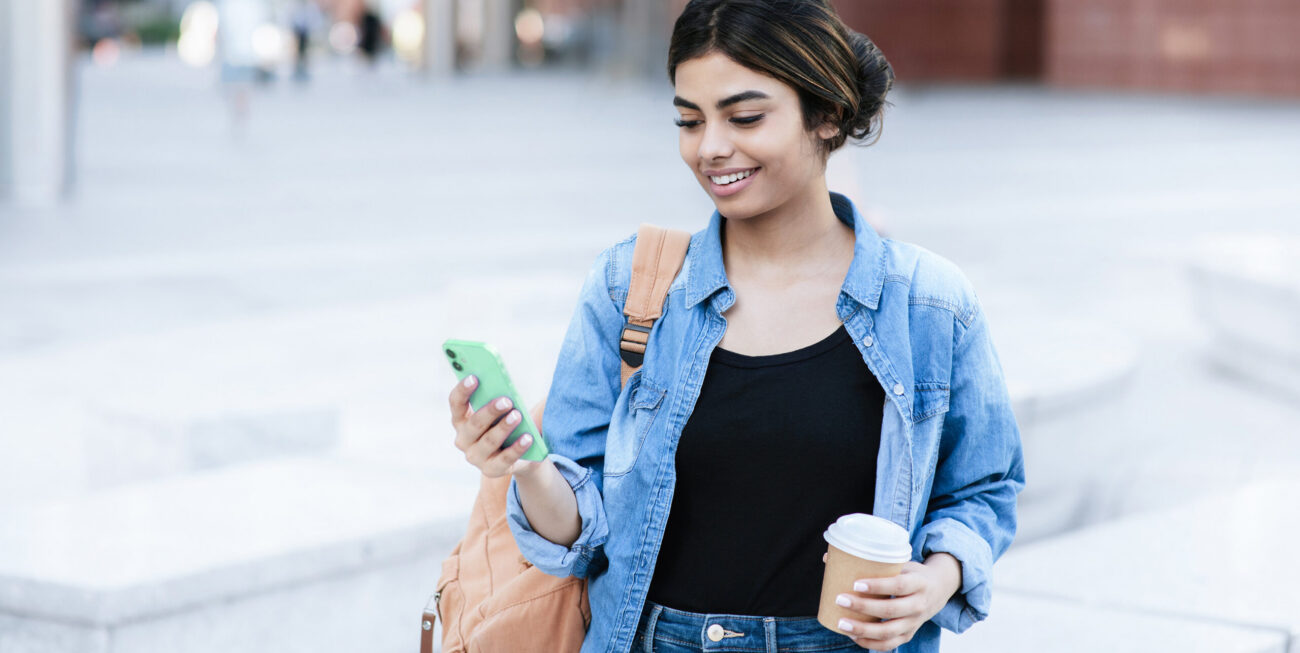 A woman looking at their cell phone with a coffee cup in their opposite hand