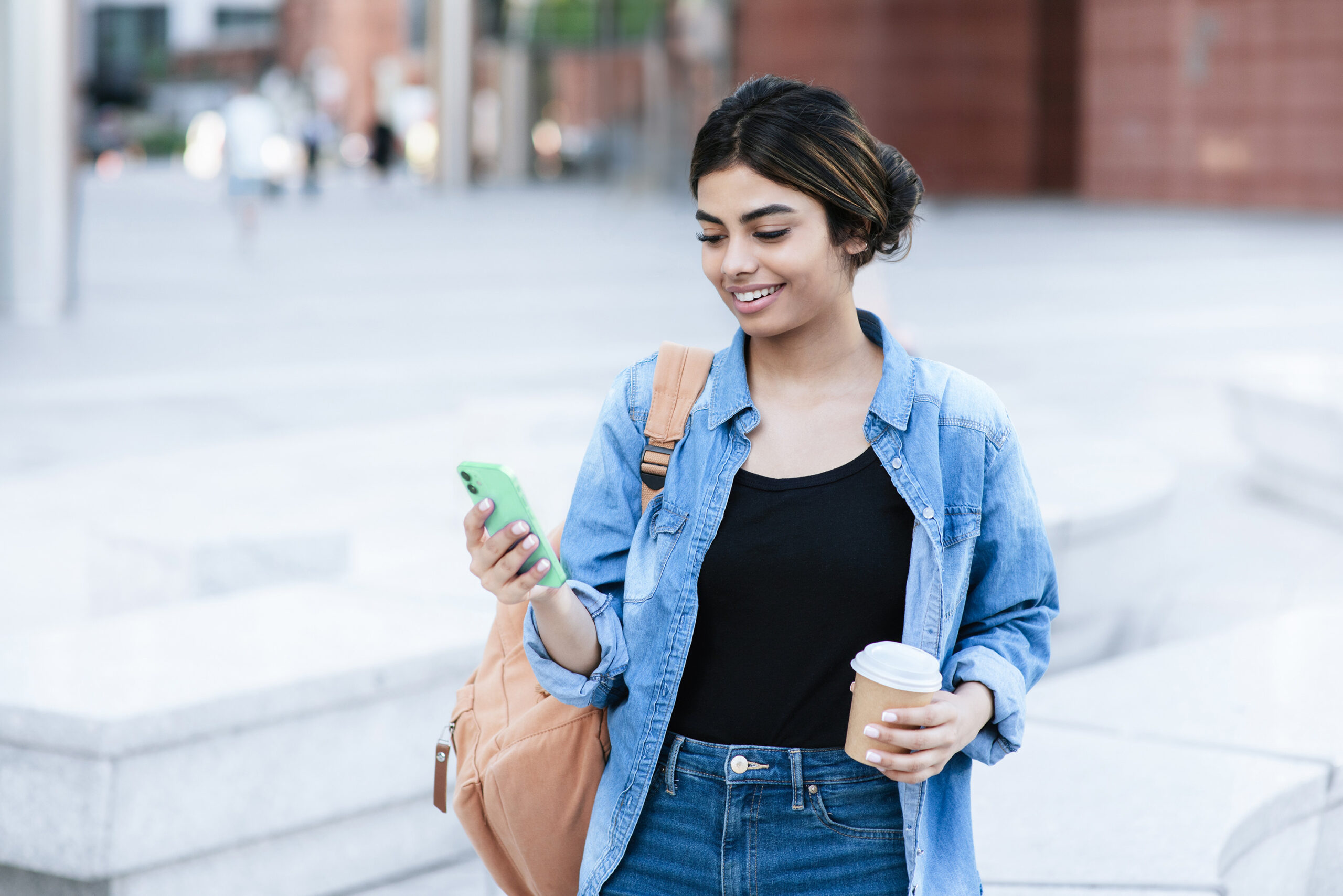 A woman looking at their cell phone with a coffee cup in their opposite hand