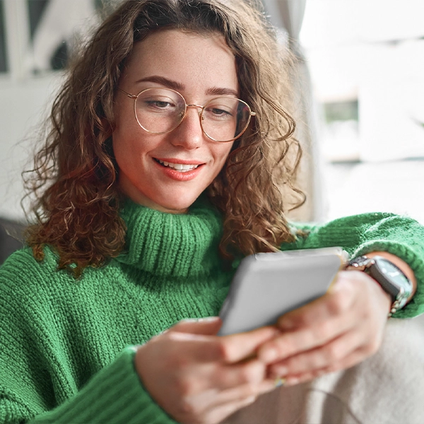 A woman with curly hair and glasses, wearing a green sweater, smiles while looking at her smartphone.