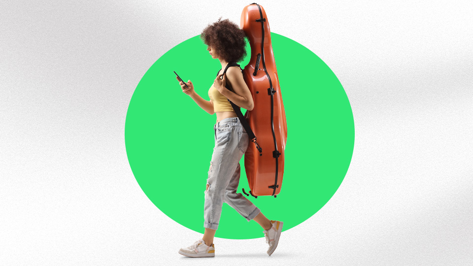 A woman walking with their phone in their hand, carrying a cello case on their back.