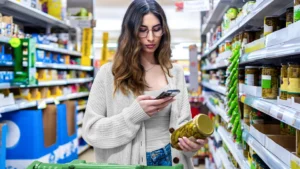 A woman looking at her cell phone in one hand and a jar of olives in the other hand.
