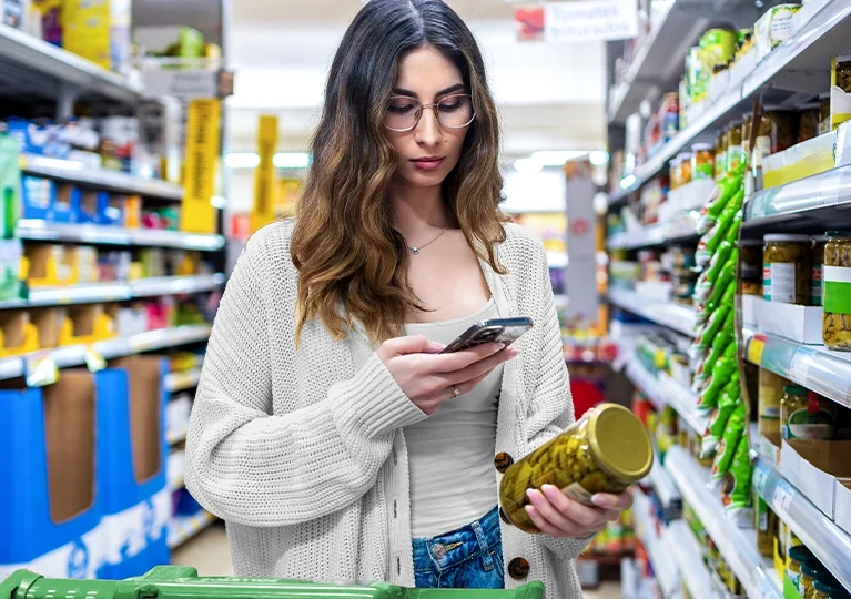 A woman looking at her cell phone in one hand and a jar of olives in the other hand.